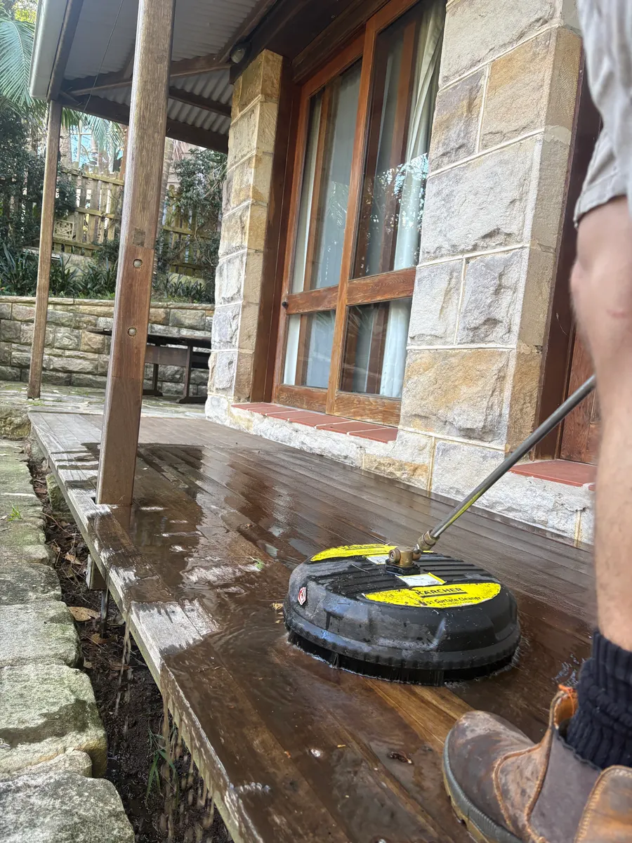 Timber deck being cleaned with a surface cleaner beside a sandstone house